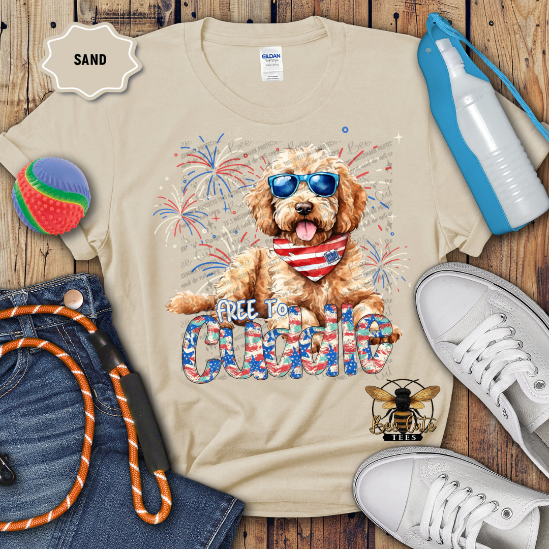 A beige colored t-shirt with a graphic print of a golden doodle dog wearing sunglasses and a patriotic 'FREE TO Cuddle' text, accompanied by Fourth of July firework graphics. The shirt is displayed flat with casual clothing items and accessories around it.