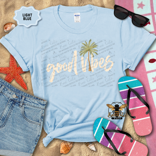 Light blue t-shirt with 'good vibes' and palm tree design, surrounded by beach accessories on sand.