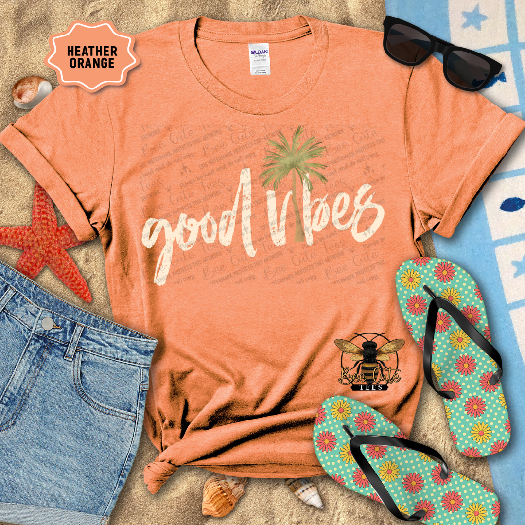 Orange t-shirt with 'good vibes' text and palm tree graphic, surrounded by beach accessories on a sandy background.