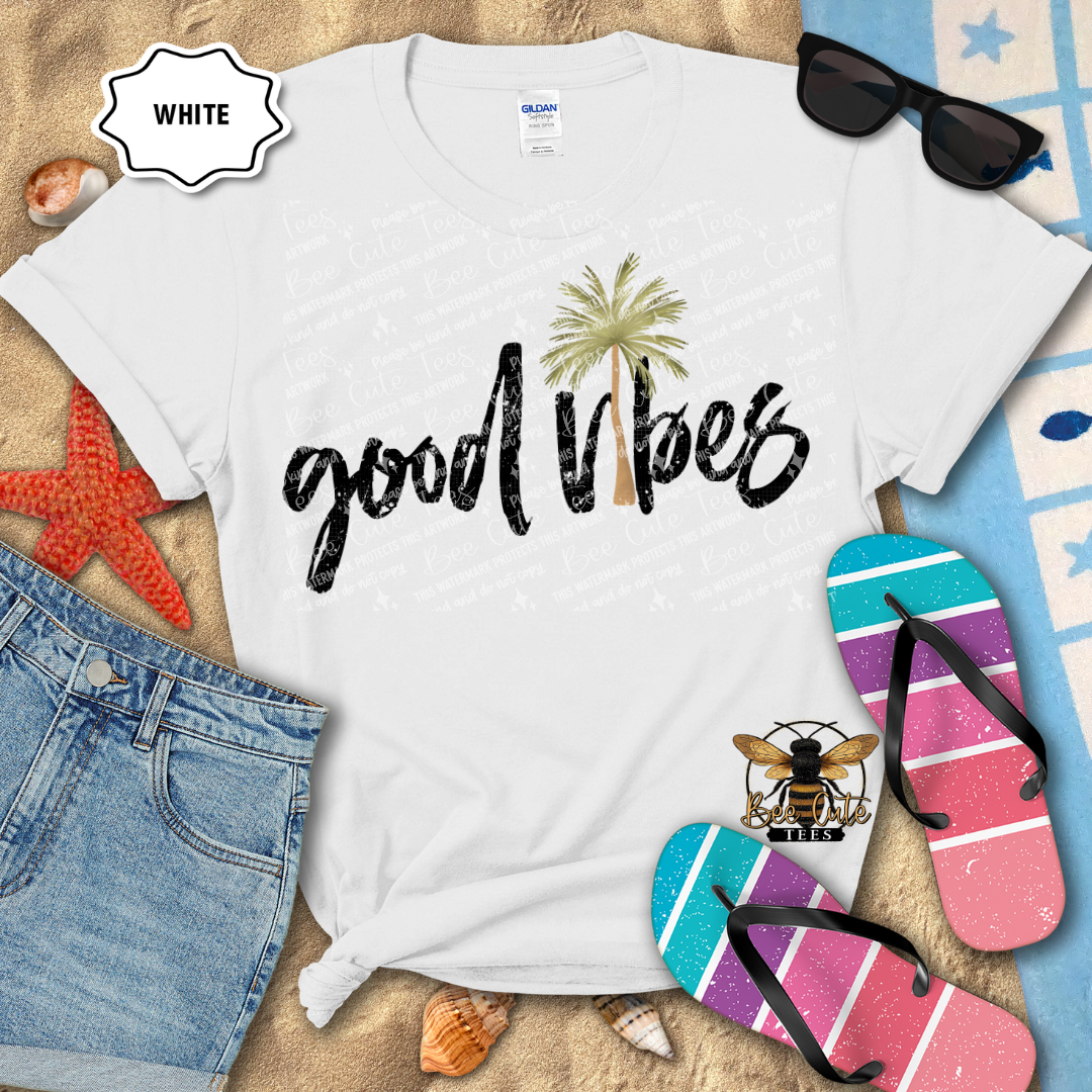 White t-shirt with 'good vibes' text and palm tree, surrounded by beach accessories on a sandy background.