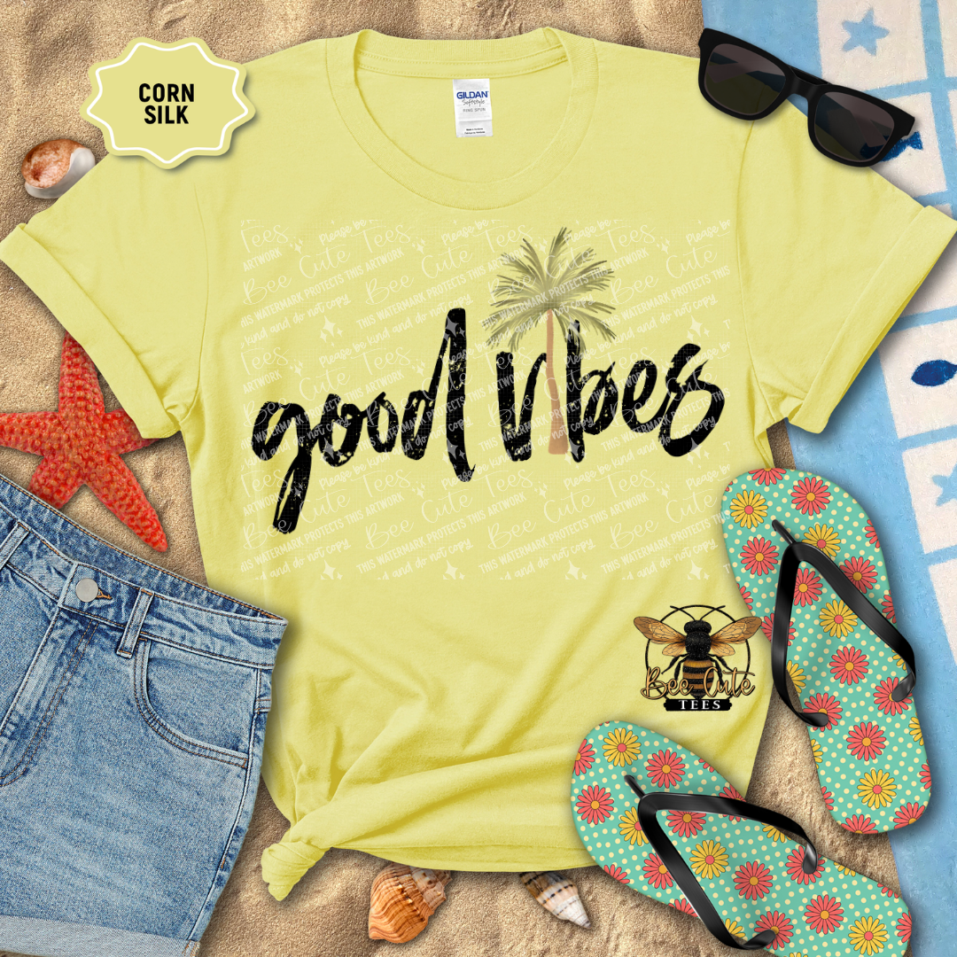 Yellow t-shirt with 'good vibes' text and palm trees, surrounded by beach items on sand.