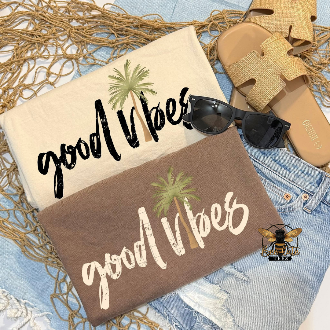 Two 'good vibes' t-shirts with sunglasses and sandals on a textured surface.