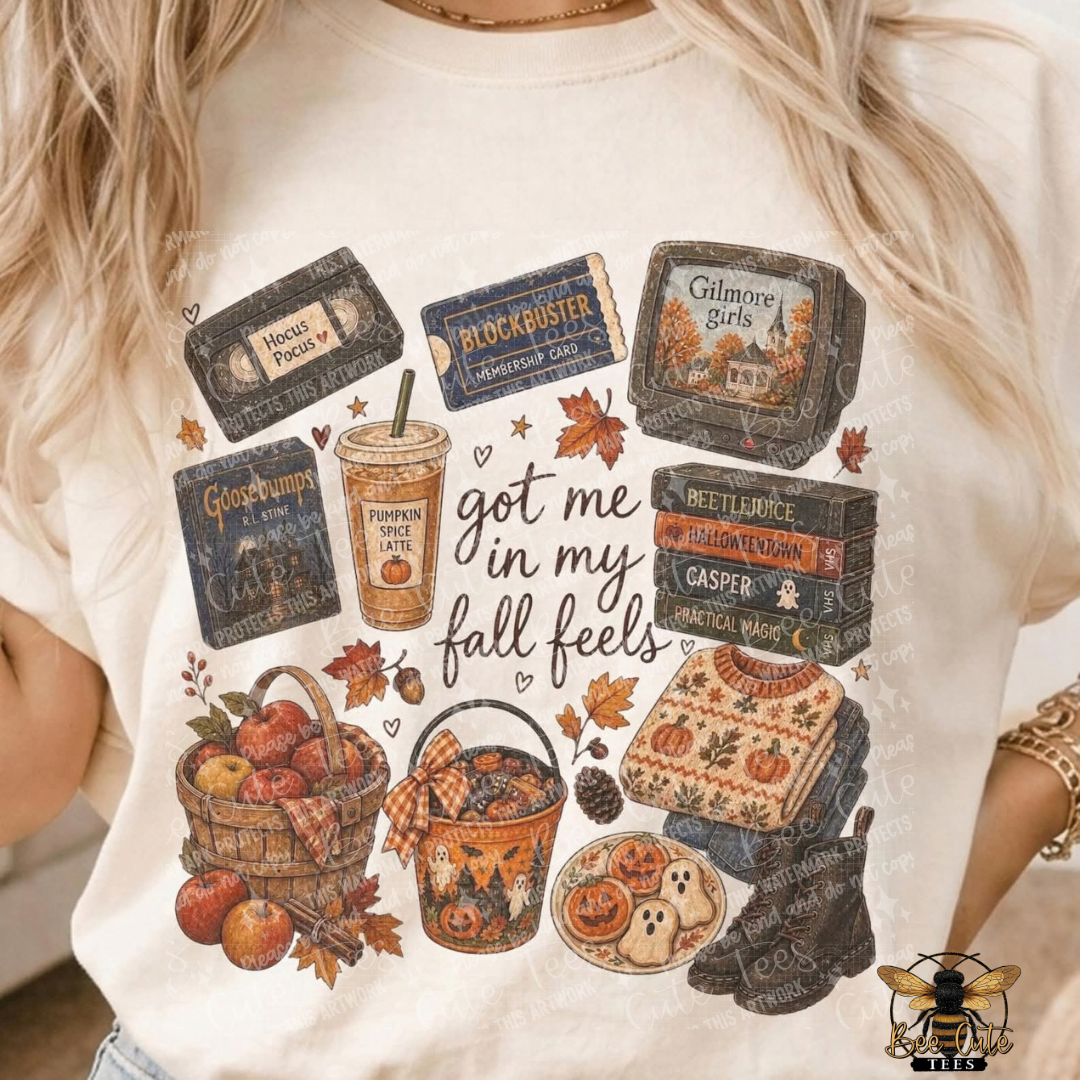 A close-up of a person wearing a cream-colored graphic t-shirt with a fall-themed design. The design features vintage book covers, a pumpkin spice latte, a basket of apples, a bucket of cookies, and autumn leaves with the text 'got me in my fall feels'.