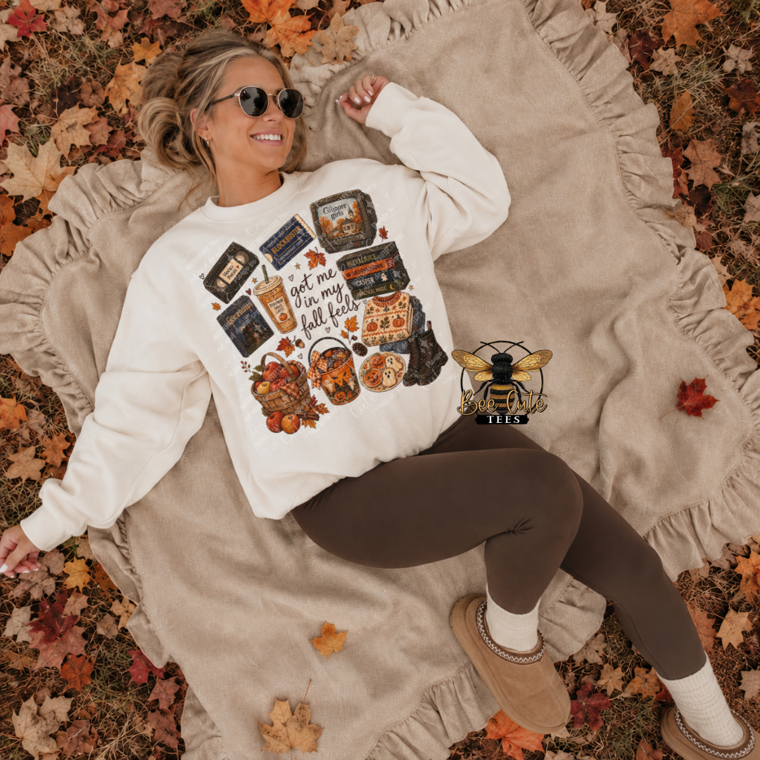A woman lying on a beige blanket outdoors among fallen autumn leaves, wearing a cream sweatshirt with a colorful fall-themed graphic and brown leggings.