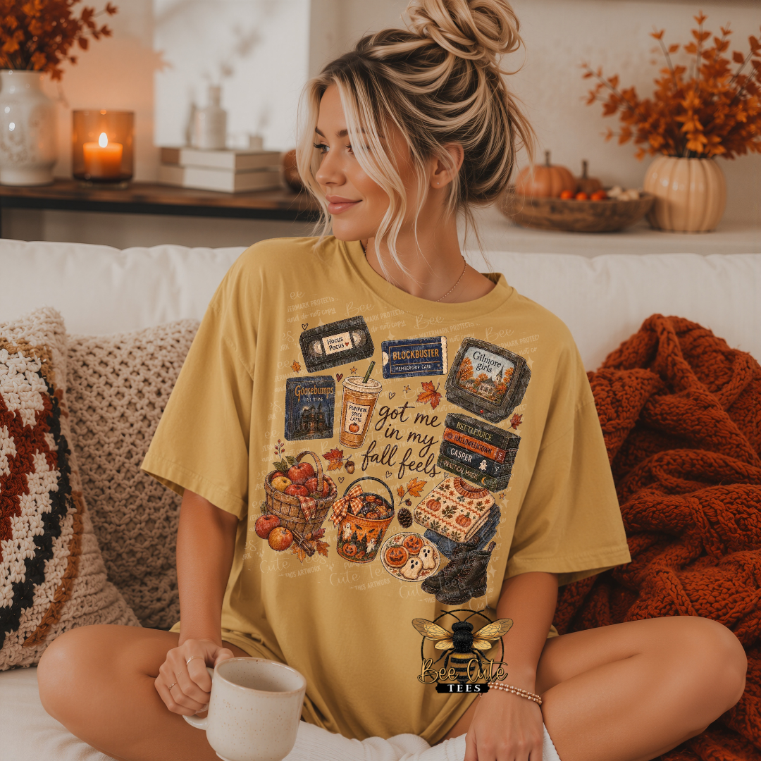 A woman sits cross-legged on a white couch wearing a mustard yellow graphic t-shirt with a fall-themed collage print. The print includes vintage cassette tapes, a coffee cup with the text 'got me in my fall falls', and autumn illustrations.