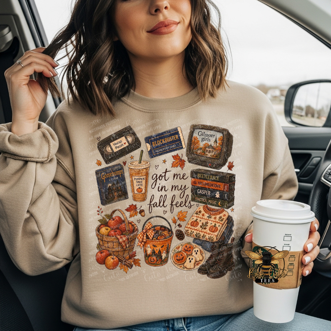 A woman sitting in a car wearing a beige crewneck sweatshirt with a detailed autumn-themed graphic print. The graphic features vintage VHS tapes, a coffee cup, fall leaves, a basket of apples, and a bucket of cookies with the text 'got me in my fall feels'.