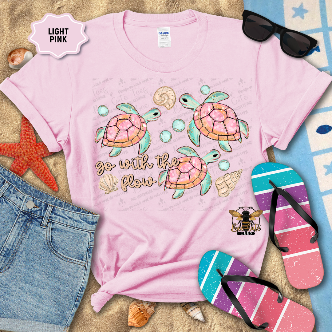 Light pink t-shirt with turtle design and 'go with the flow' text, surrounded by beach accessories.