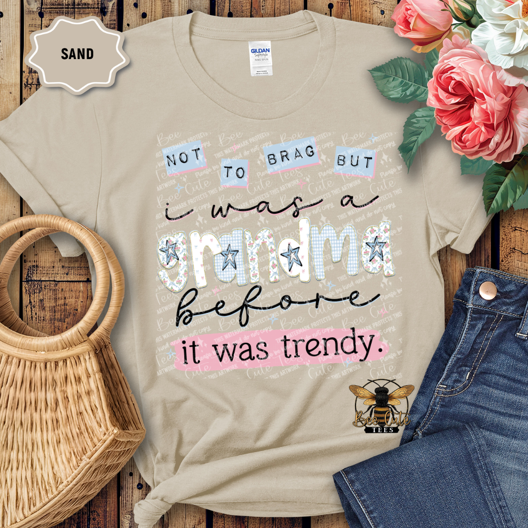 Beige t-shirt with text 'Not to brag but I was a grandma before it was trendy' on a wooden surface with flowers and denim jeans.