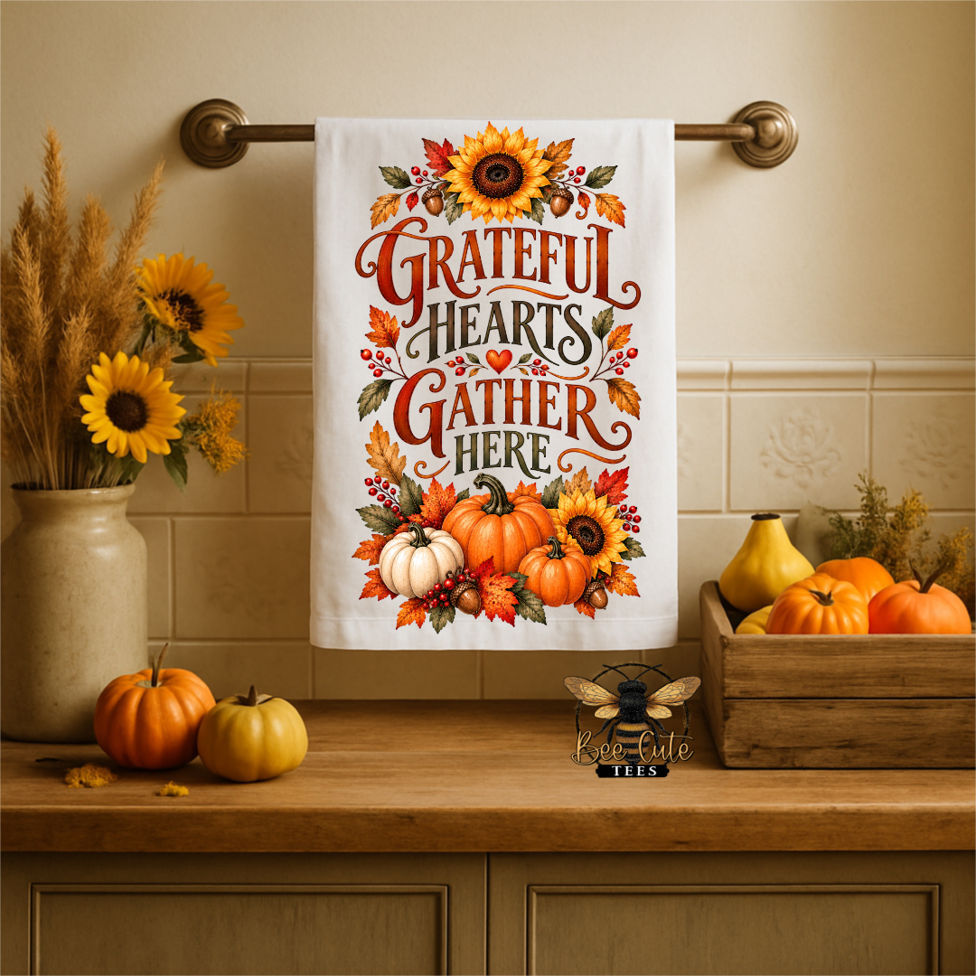 A white kitchen towel with a colorful autumn design hangs from a brass rack on a beige tiled wall above a wooden counter. The design features the phrase 'Grateful Hearts Gather Here' in stylized lettering, surrounded by sunflowers, pumpkins, and fall leaves.