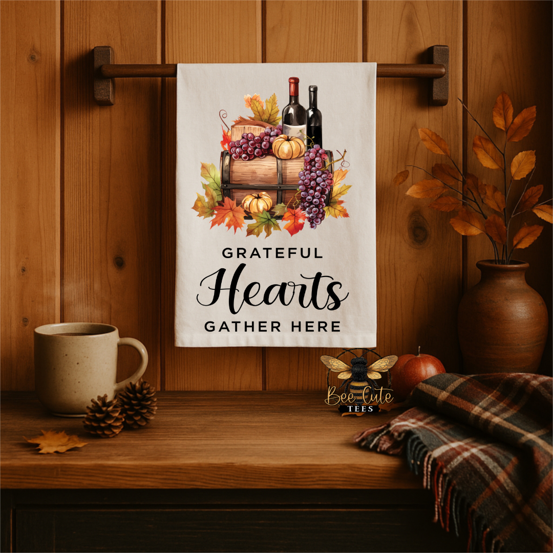 A white kitchen towel with an autumn harvest illustration and the text 'GRATEFUL Hearts GATHER HERE' hanging on a wooden wall rack in a cozy, rustic kitchen setting.