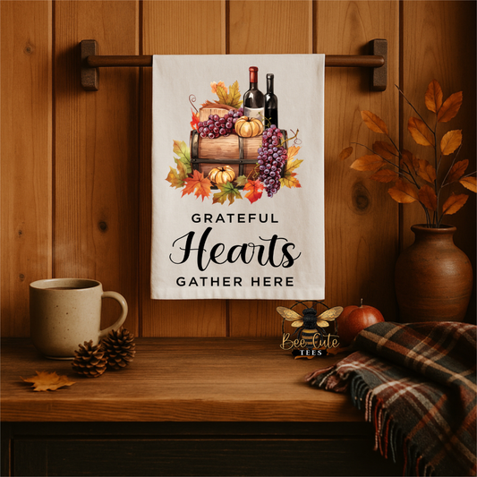 A white kitchen towel with an autumn harvest illustration and the text 'GRATEFUL Hearts GATHER HERE' hanging on a wooden wall rack in a cozy, rustic kitchen setting.