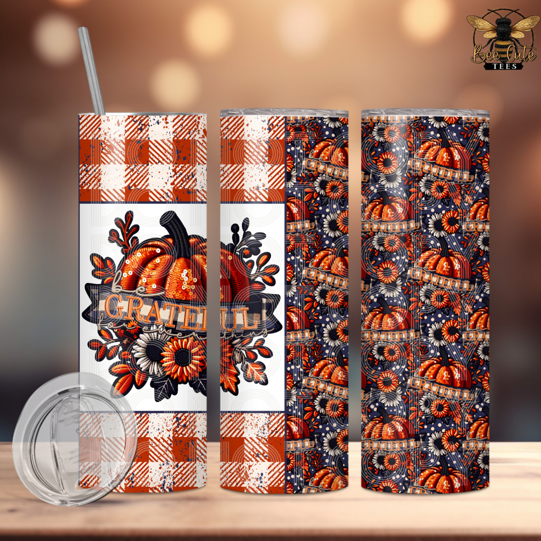 Three views of a 20 oz skinny tumbler with a Halloween-themed design, including orange and white gingham borders, a central pumpkin illustration with the word 'Grateful', and a dark navy background with orange pumpkins and white snowflakes. A clear lid is shown in the foreground on a wooden surface with a warm bokeh background.