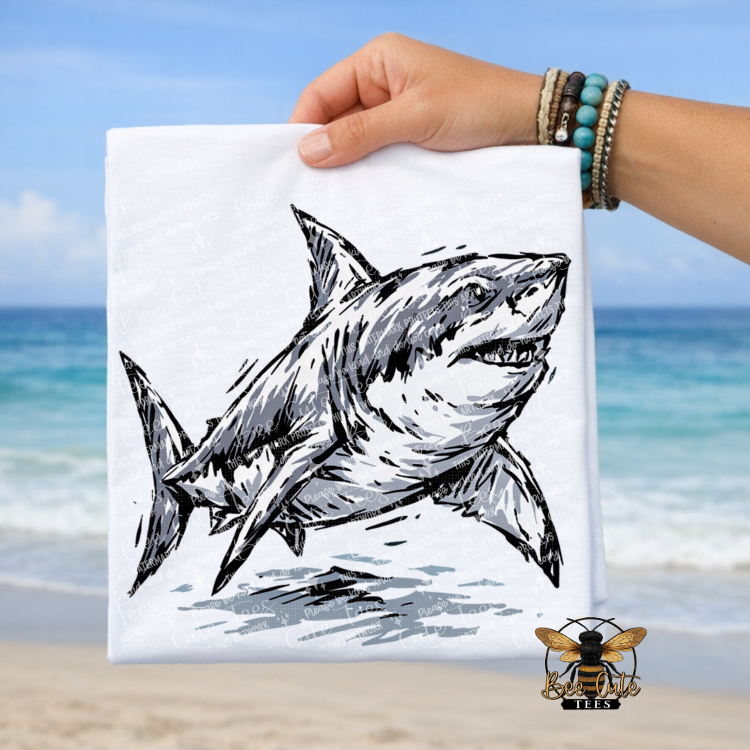 A hand holds a white folded t-shirt featuring a black-and-white shark illustration, with a beach and ocean in the background. A bee logo appears in the lower right corner.