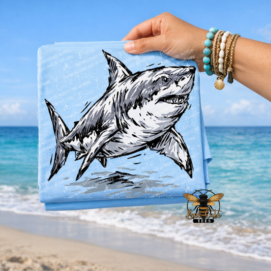 A light blue folded t-shirt held by a hand over a beach background, featuring a large black and white shark illustration. The background shows a sandy beach, turquoise ocean, and blue sky.