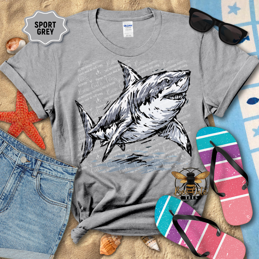 A heather gray graphic t-shirt featuring a detailed illustration of a great white shark, styled in a flat-lay arrangement with blue denim shorts, pink and blue flip-flops, sunglasses, and beach-themed props on a sandy background.