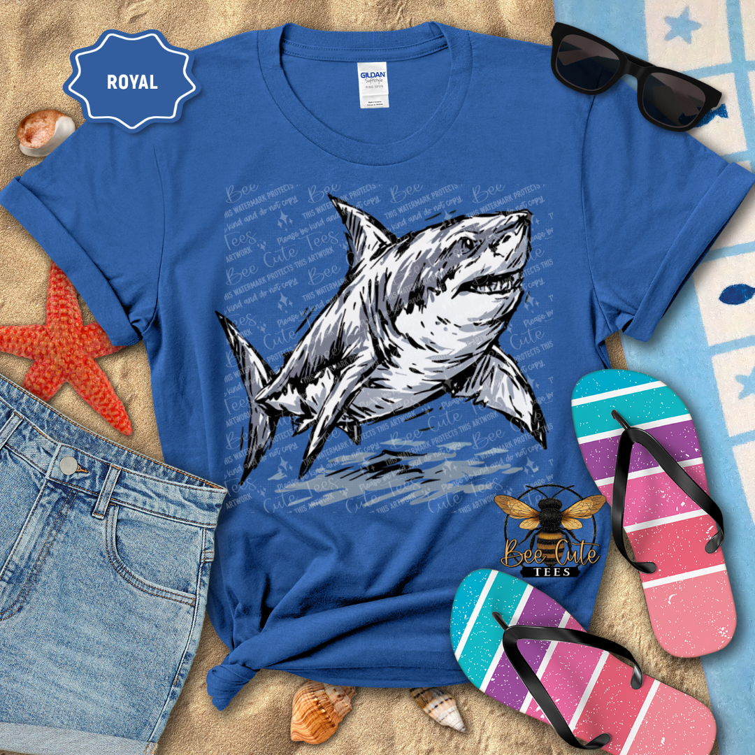 A royal blue graphic t-shirt featuring a detailed illustration of a great white shark swimming over nautical chart text. The shirt is styled in a beach-themed flat lay with blue denim shorts, pink and teal striped flip-flops, black sunglasses, a red starfish, and seashells on a sandy background.