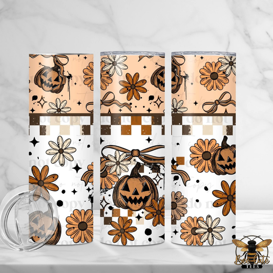 Three slim stainless steel tumblers with a Halloween-themed pattern featuring jack-o'-lanterns, daisies, bows, stars, and checkerboard accents in orange, brown, beige, and white. A clear lid is shown in the lower left on a light marble surface.