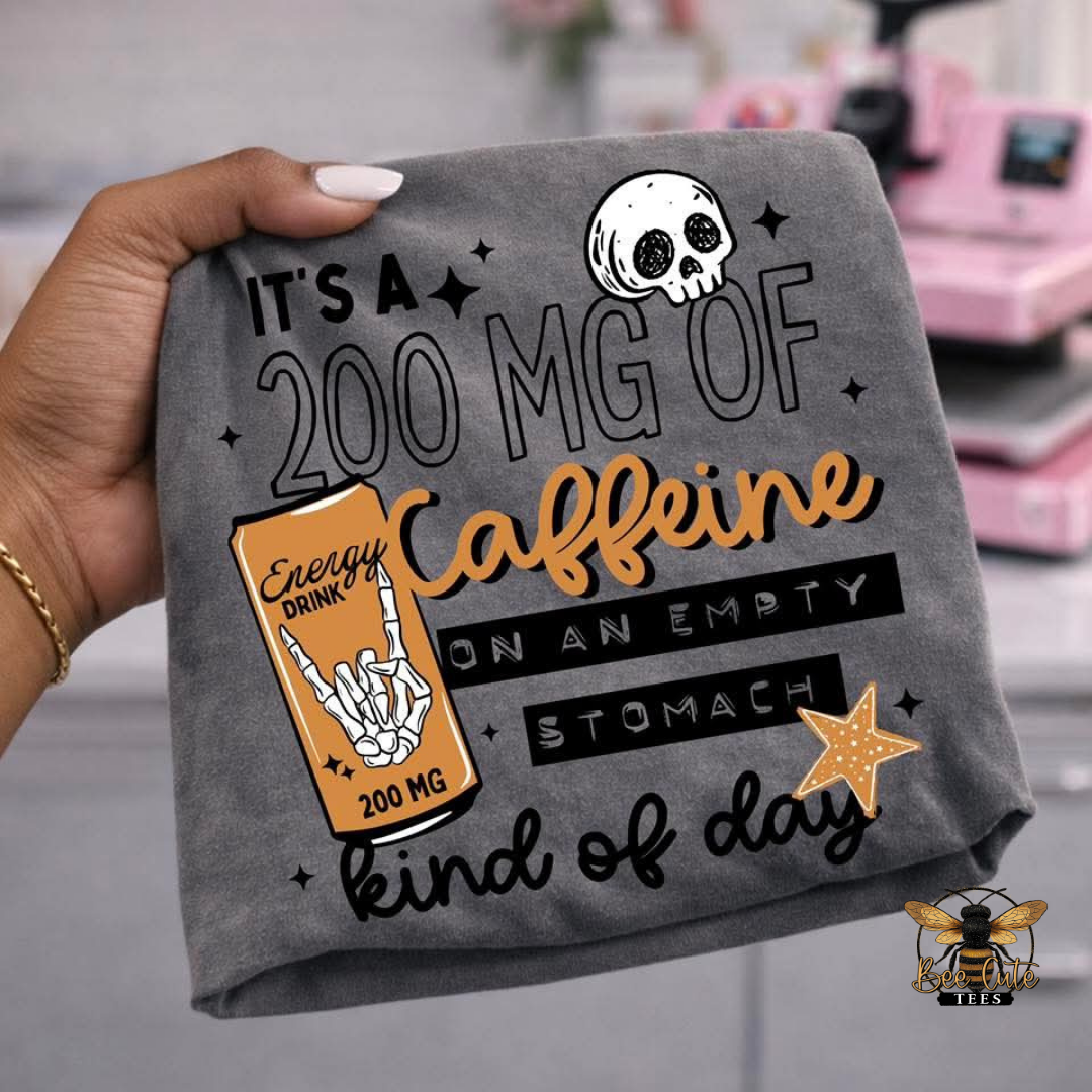 A hand holds a dark gray t-shirt featuring a humorous graphic that reads 'IT'S A 200 MG OF Caffeine ON AN EMPTY STOMACH kind of day' with a skull, a coffee can, and a star graphic.
