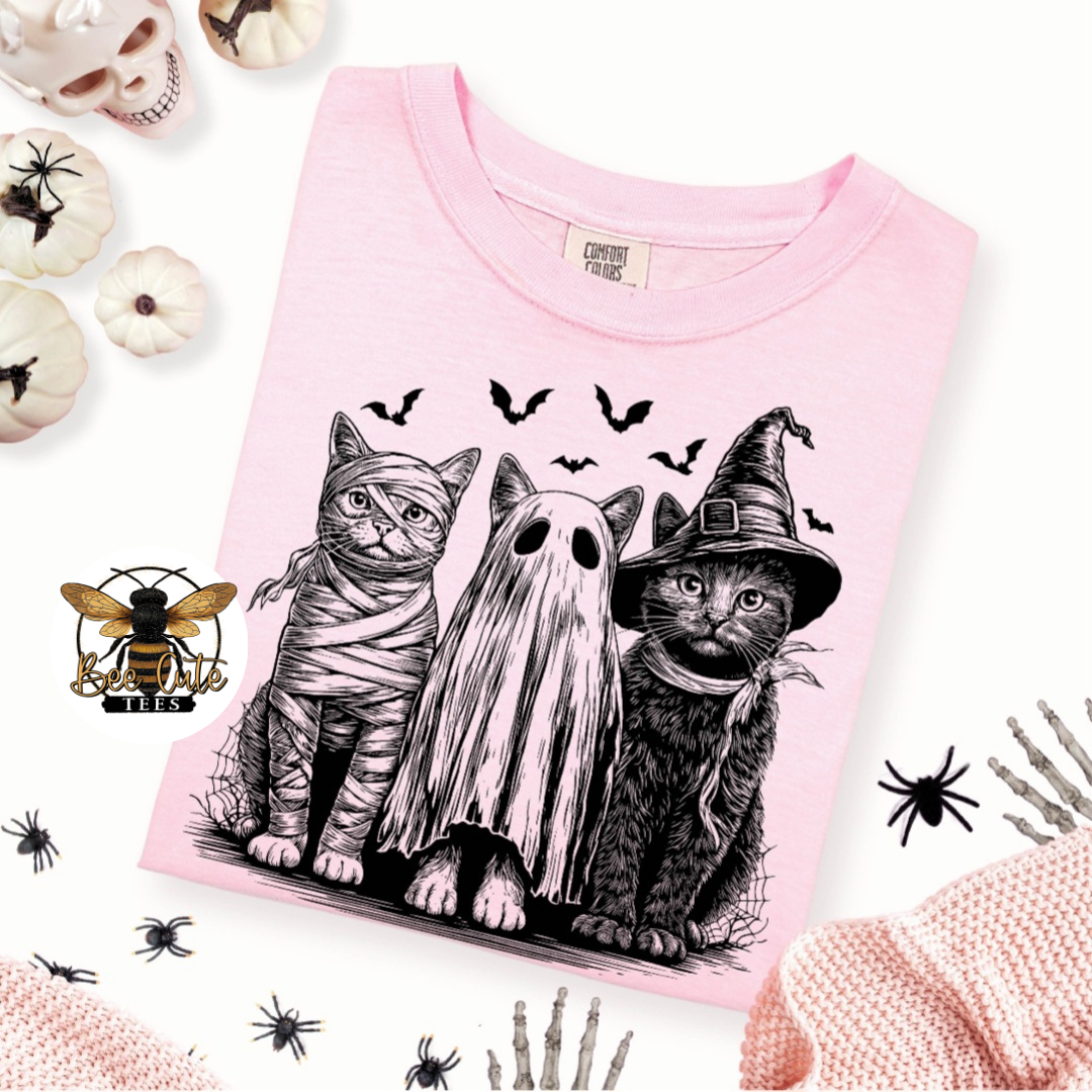 A pink short-sleeve T-shirt laid flat on a white background, featuring a black line-art graphic of three cats dressed as Halloween characters: a mummy, a ghost, and a witch. The shirt is styled with Halloween props including pumpkins, spiders, bones, and a bee logo.