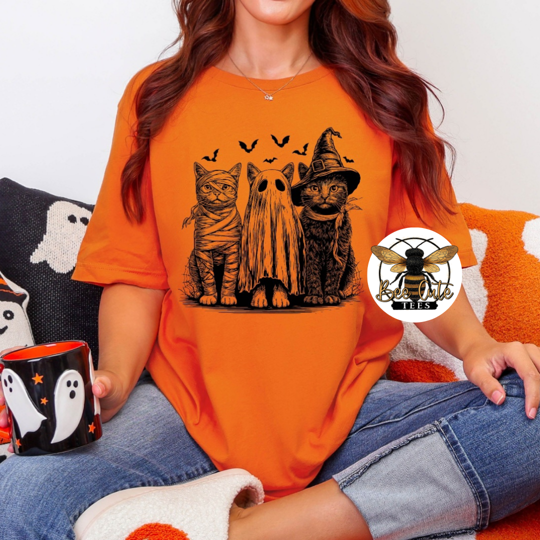 A woman wearing an orange graphic t-shirt with a black print of three cats dressed as mummies, a ghost, and a witch, sitting on a couch and holding a black mug with a ghost design.
