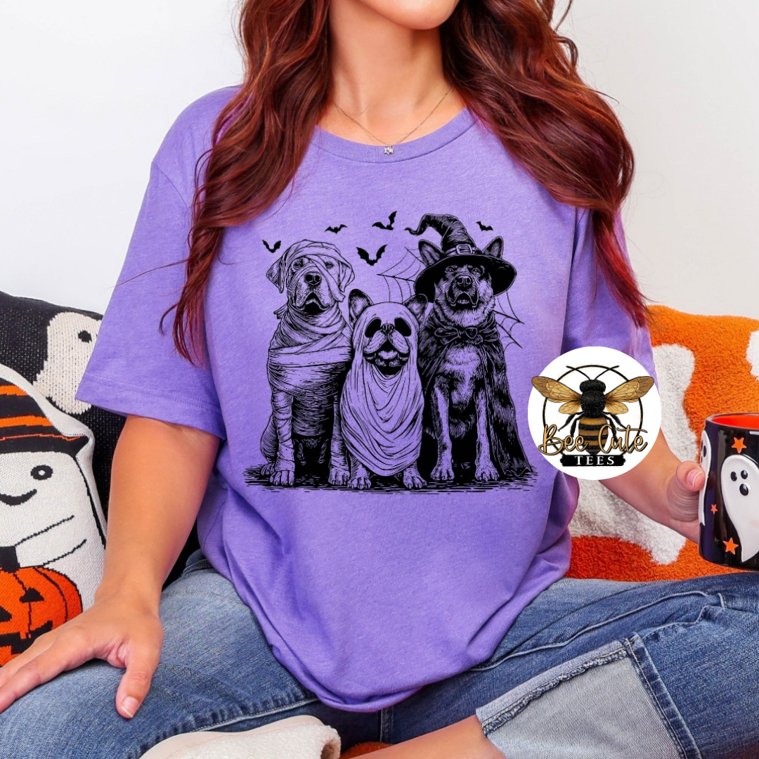 A woman wearing a purple graphic t-shirt with a black and white print of three dogs dressed as Halloween characters (wrapped in bandages, in a witch hat, and standing). She is sitting on a couch holding a mug.