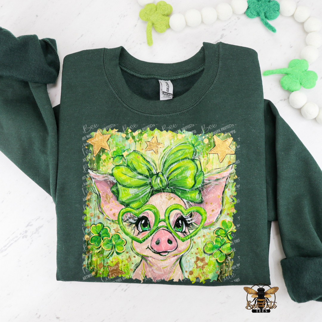 Green sweatshirt with a pig design wearing glasses and a bow, surrounded by shamrocks on a white background.