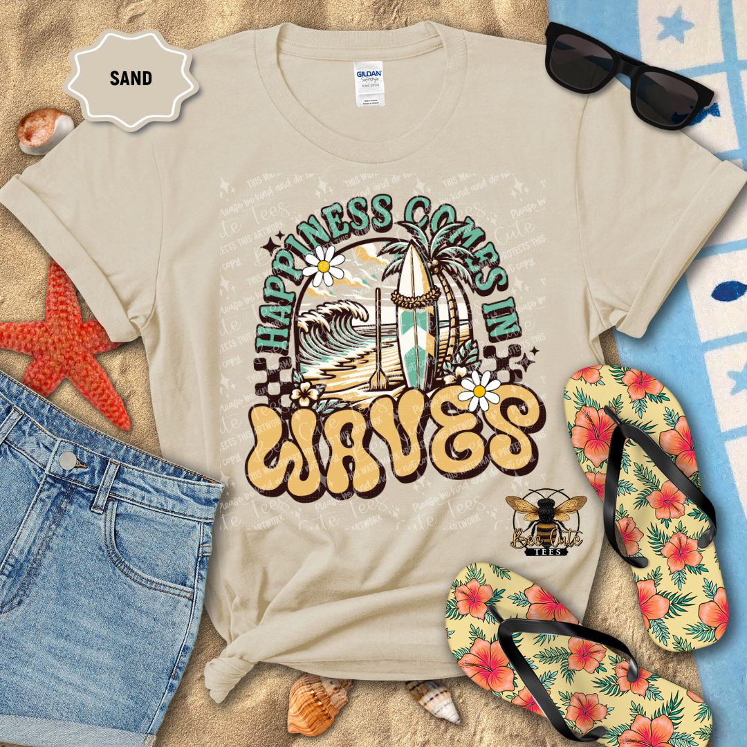 Beige t-shirt with 'Happiness Comes in Waves' design on sand with beach accessories.