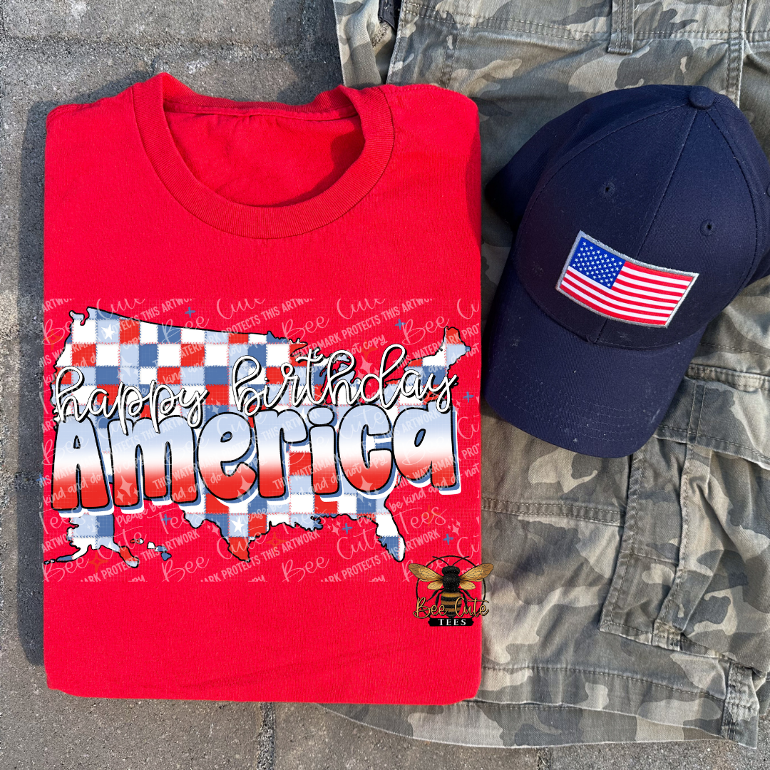 Red t-shirt with 'Happy Birthday America' design, navy cap with American flag, and camouflage pants on a textured surface.