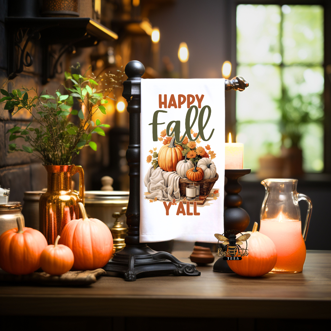 A decorative towel-style sign on a black stand, printed with the phrase “Happy Fall Y’All” and an autumn harvest illustration with pumpkins, gourds, leaves, and a basket. The sign is staged on a wooden table in a warm, dark room with candles, pumpkins, a pitcher, and soft window light in the background.