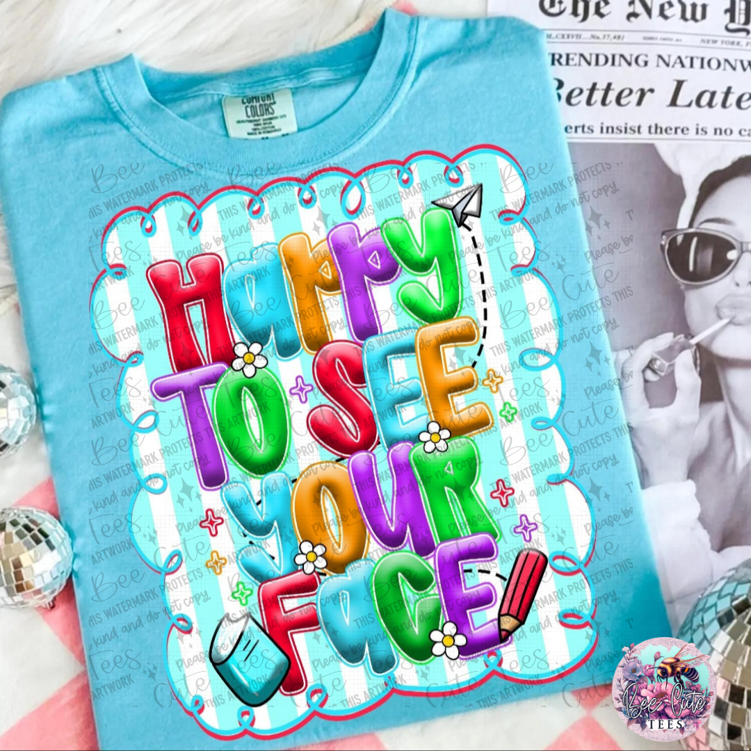 Blue t-shirt with colorful text 'Happy to see your face!' on a surface with a magazine in the background.