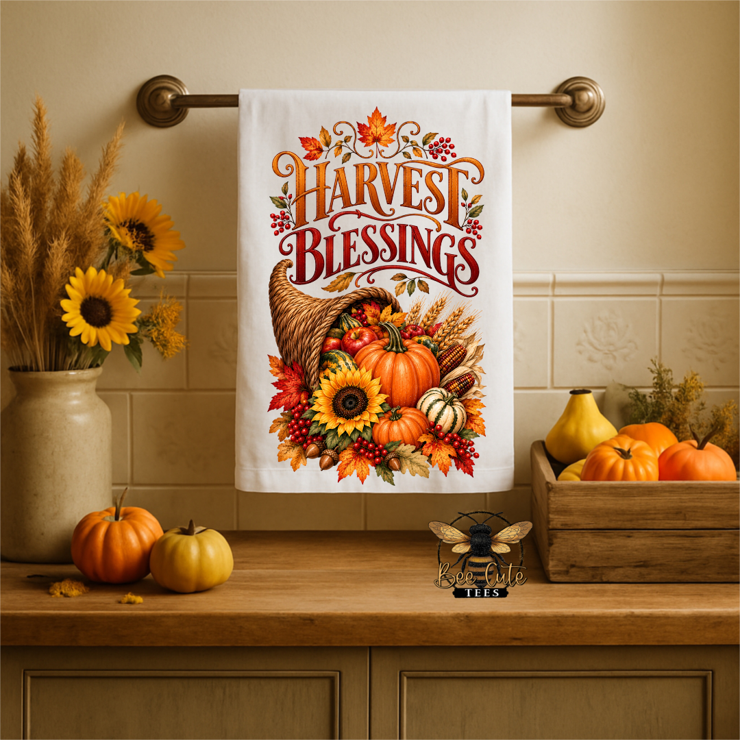 A white kitchen towel with a colorful 'Harvest Blessings' autumn print hangs on a brass towel bar over a wooden countertop. The scene is styled with fall decorations including sunflowers, pumpkins, and a wooden crate.