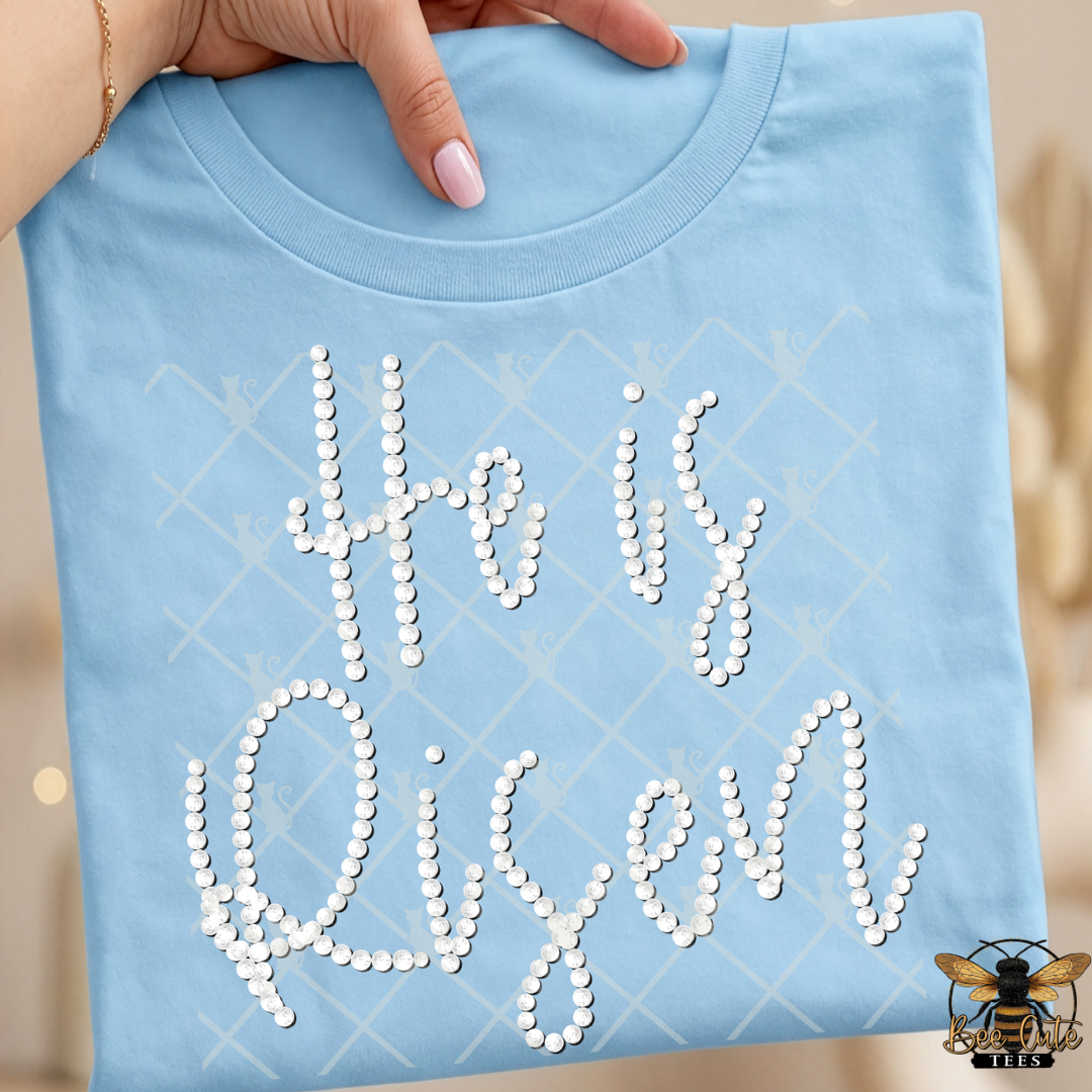 Light blue t-shirt with 'He is Risen' text made of faux white pearls, held by a hand.