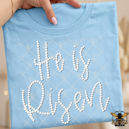 Light blue t-shirt with 'He is Risen' text made of faux white pearls, held by a hand.