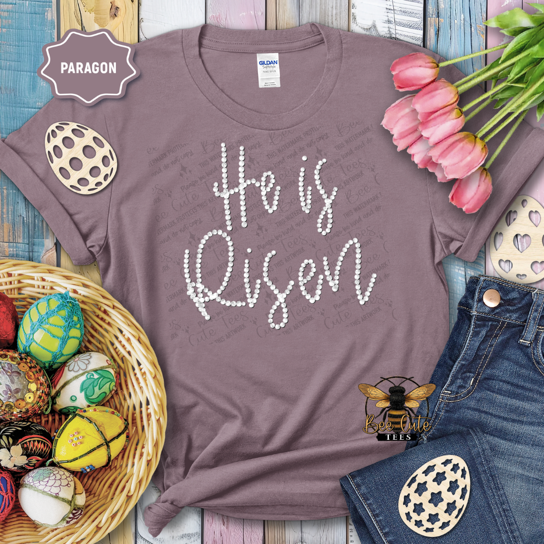 Purple t-shirt with 'He is Risen' text, surrounded by Easter decorations on a wooden surface.