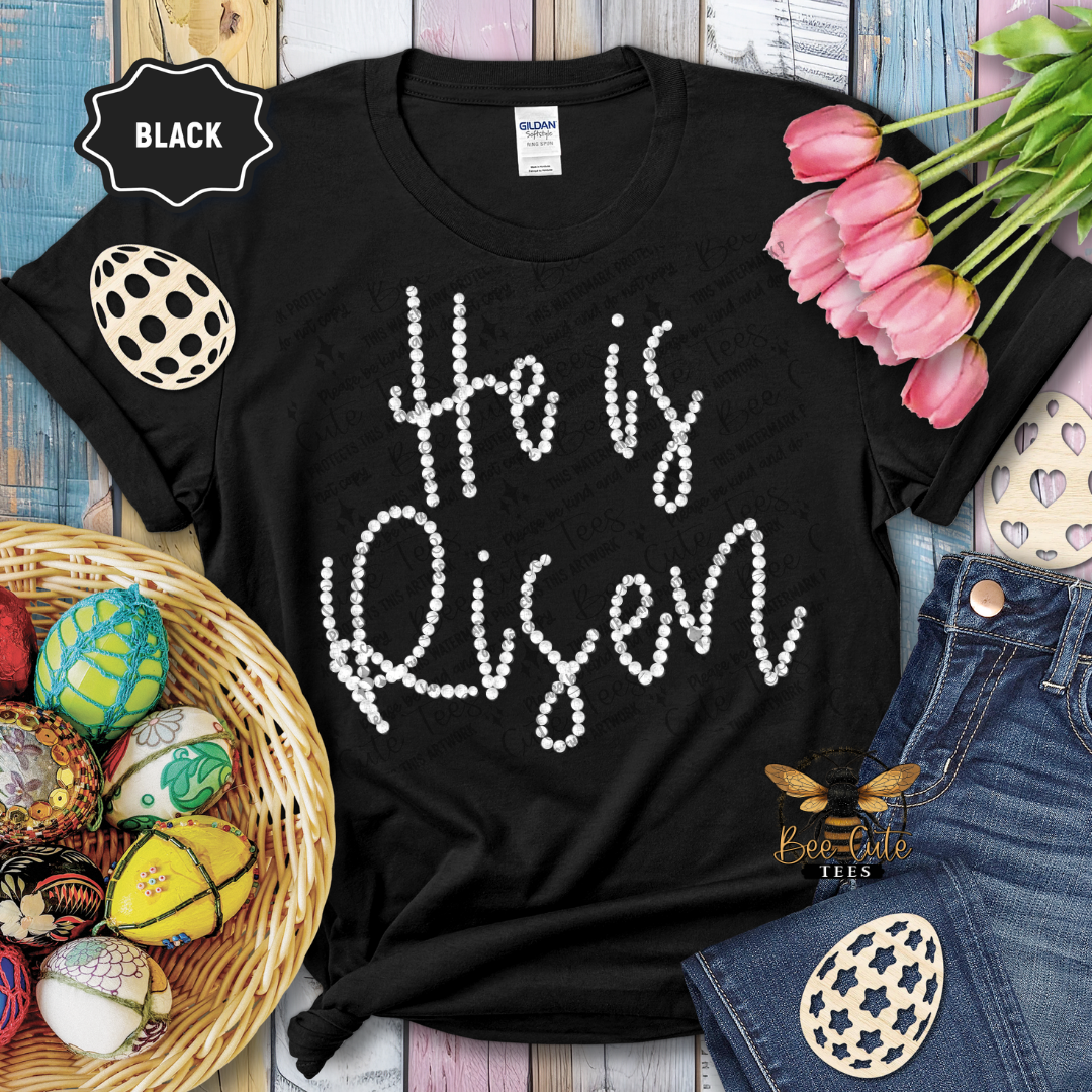 Black t-shirt with 'He is Risen' text, Easter eggs, and tulips on a wooden surface.
