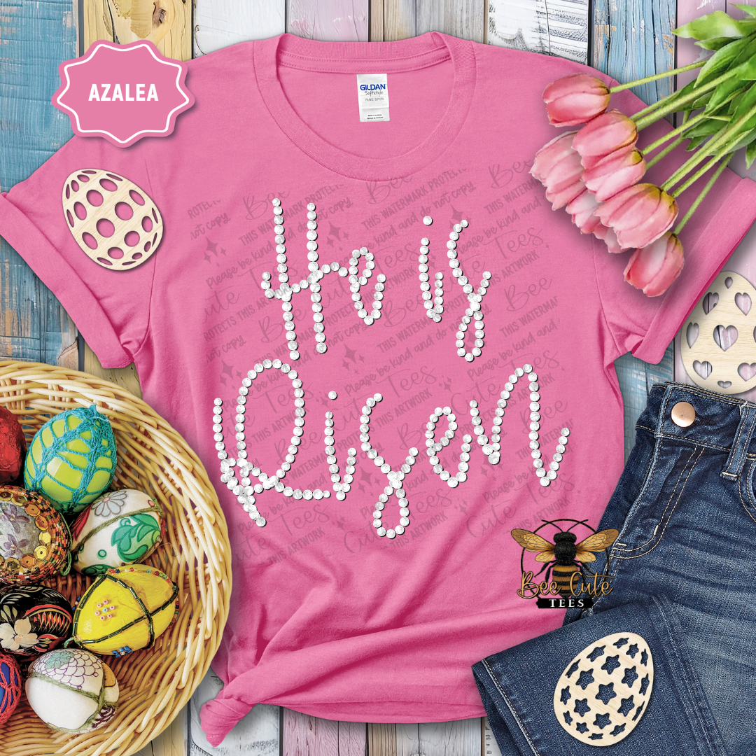 Pink t-shirt with 'He is Risen' text, surrounded by Easter decorations on a wooden surface.