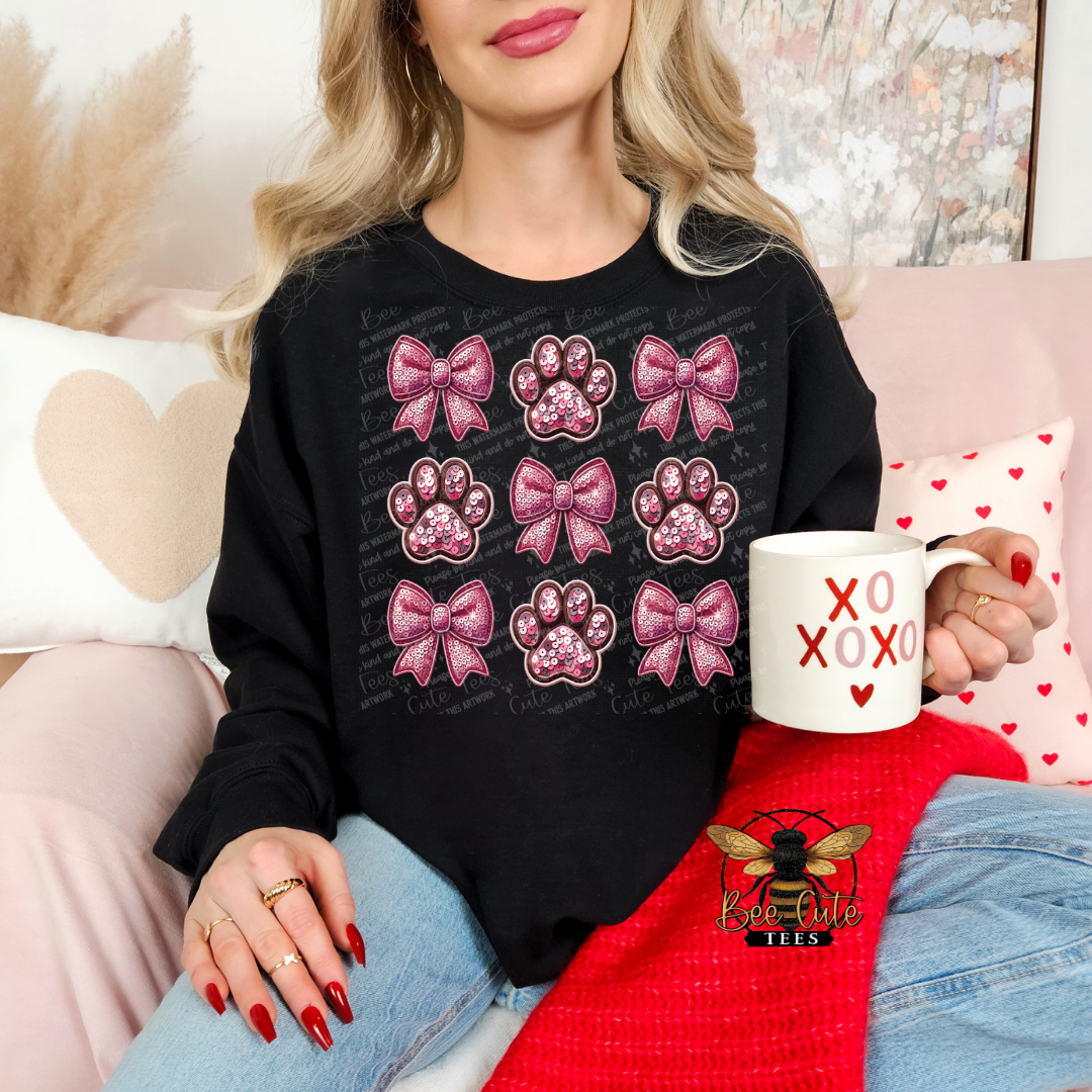 Person wearing a black sweatshirt with pink bows and paw prints, holding a mug with 'XOXO' text, sitting on a couch.