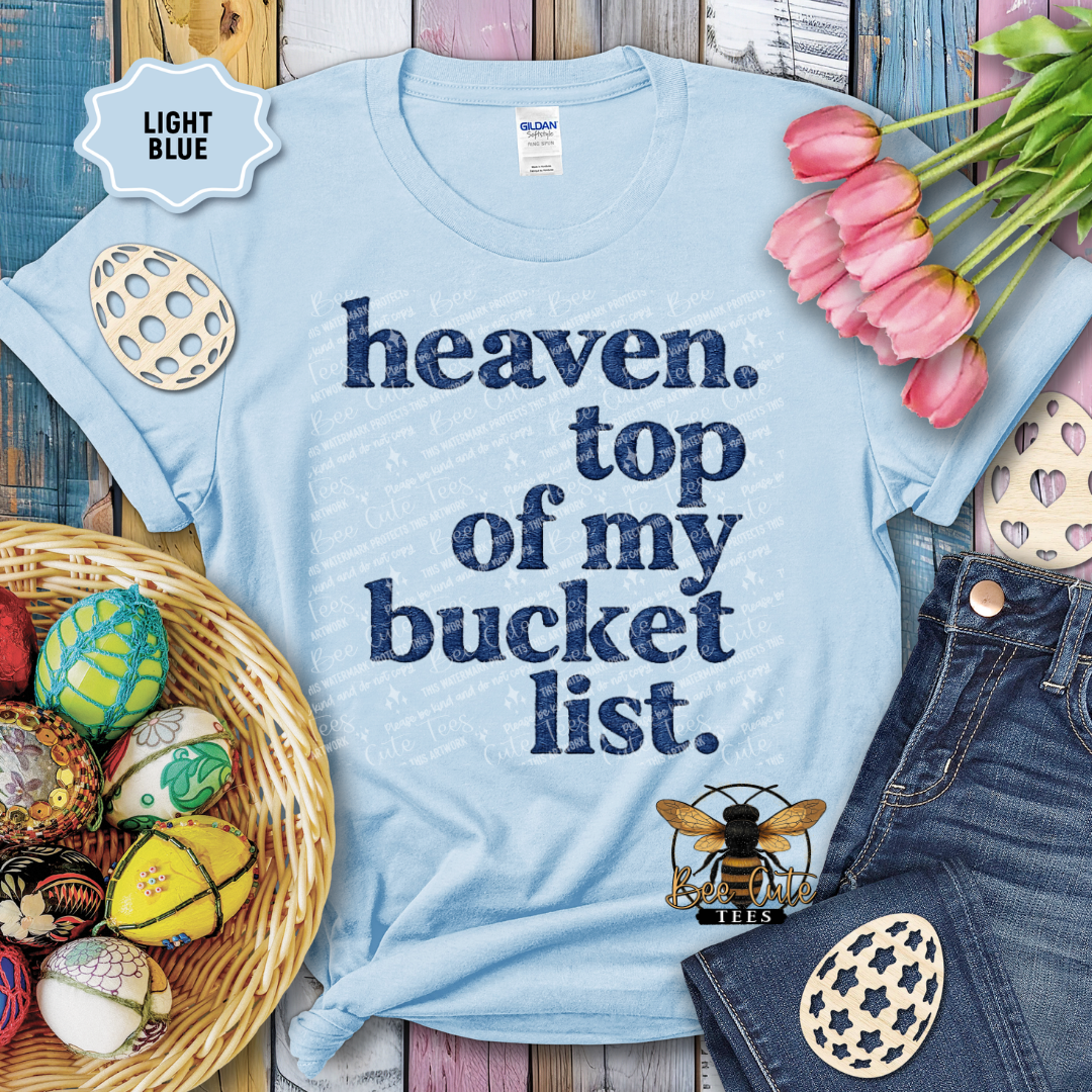 Light blue t-shirt with 'heaven. top of my bucket list.' text, surrounded by decorative items on a wooden surface.