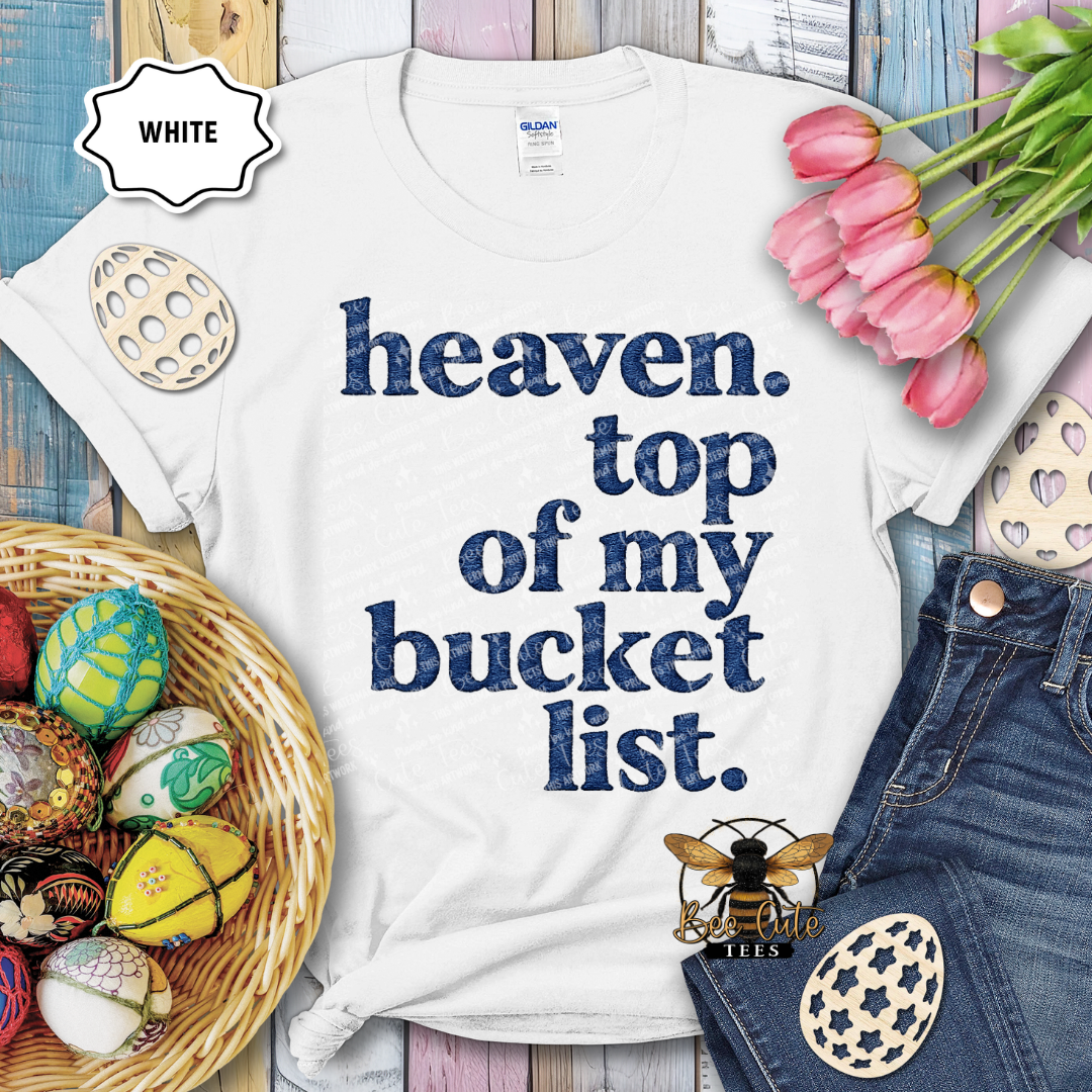 White t-shirt with 'heaven. top of my bucket list.' text, surrounded by Easter decorations on a wooden surface.