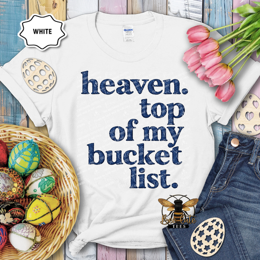 White t-shirt with 'heaven. top of my bucket list.' text, surrounded by Easter decorations on a wooden surface.