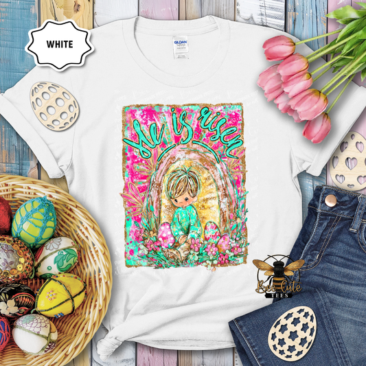 White t-shirt with 'She is Risen' design featuring a child and floral elements, surrounded by Easter decorations.