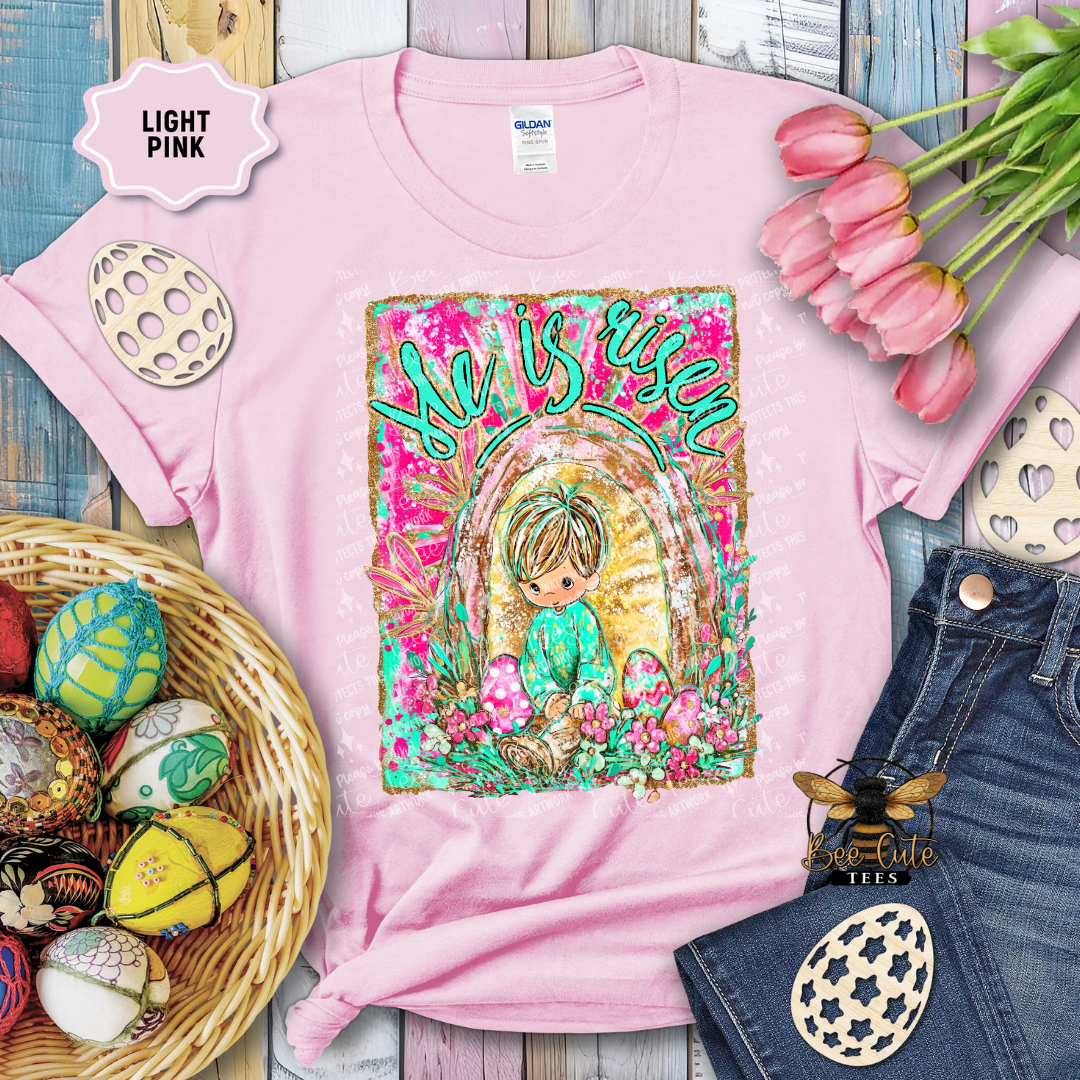 Light pink t-shirt with Easter-themed design featuring a child and 'She is risen' text, surrounded by Easter decorations.