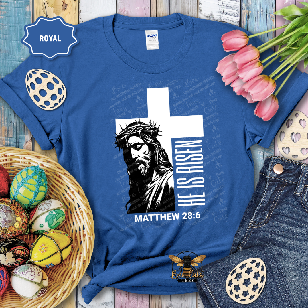 Blue t-shirt with Jesus graphic and 'He is Risen' text on a wooden surface with Easter decorations.