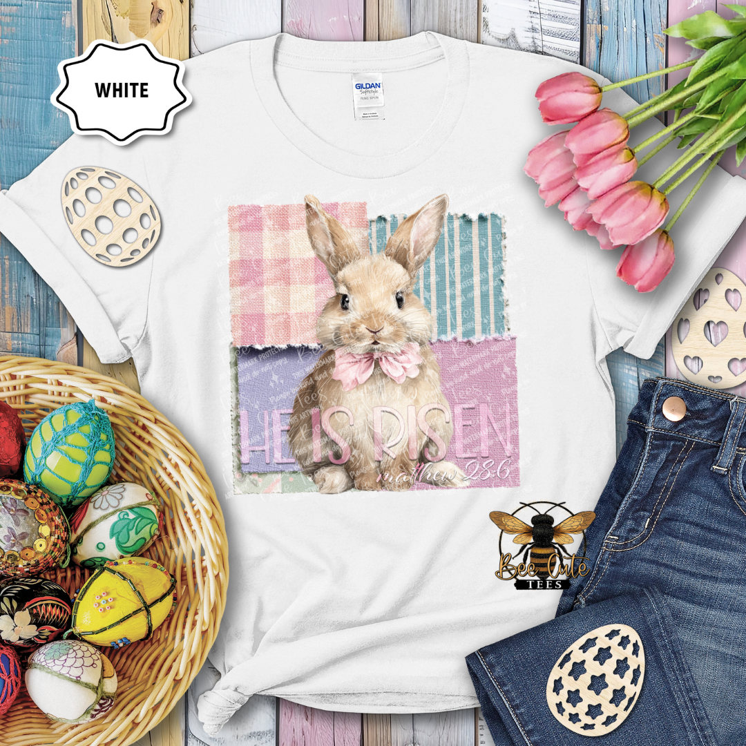 White t-shirt with a bunny graphic and 'He is risen' text, surrounded by Easter decorations.