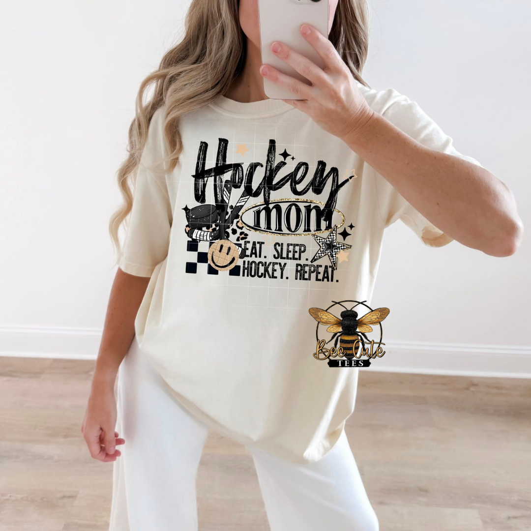 A woman takes a mirror selfie wearing a cream-colored t-shirt with a black graphic that reads 'Hockey mom' and 'Eat. Sleep.