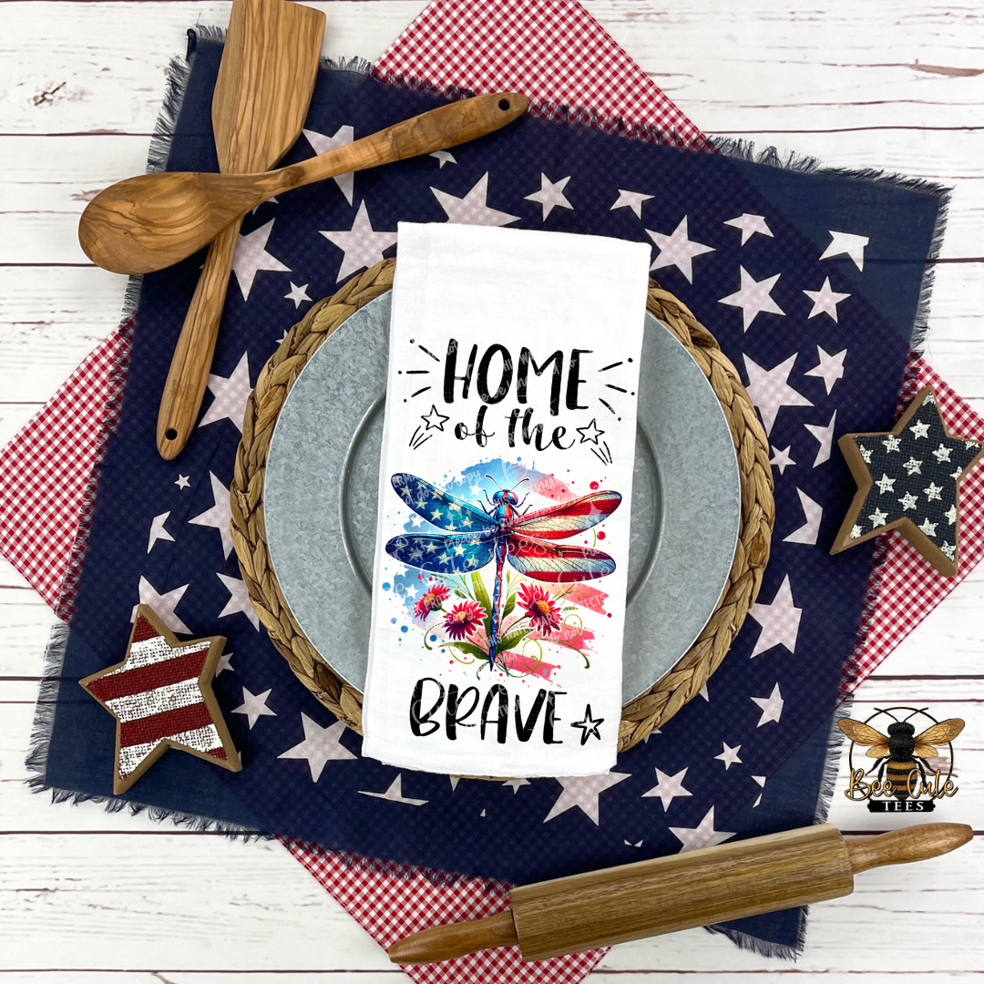 White kitchen towel displayed on a patriotic table setting, featuring a red, white, and blue dragonfly with flowers and the phrase “Home of the Brave,” surrounded by star-patterned linens, wooden utensils, and rustic decor.
