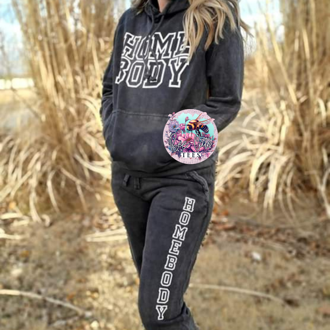 Person wearing a black 'HOMEBODY' hoodie and pants with a colorful logo on a natural background.