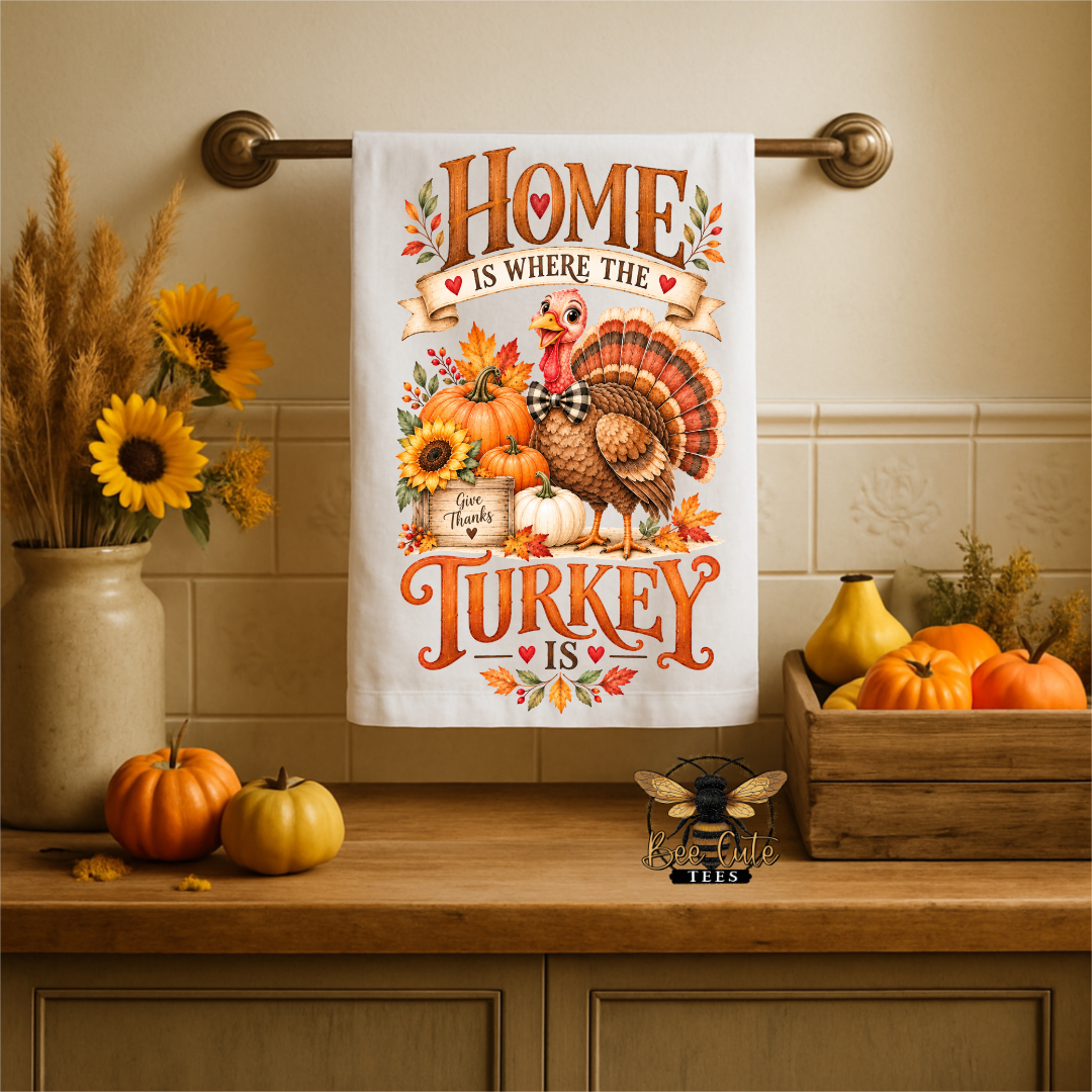 A white kitchen towel with a colorful autumn Thanksgiving design hangs on a brass towel bar. The towel features an illustration of a turkey, pumpkins, sunflowers, and the text 'HOME IS WHERE THE TURKEY IS' and 'Thanks for Sharing'.