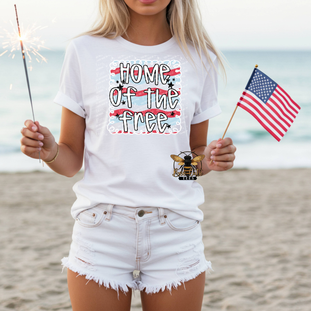 Person on a beach wearing a white t-shirt with 'Home of the Free' design, holding an American flag and a sparkler.