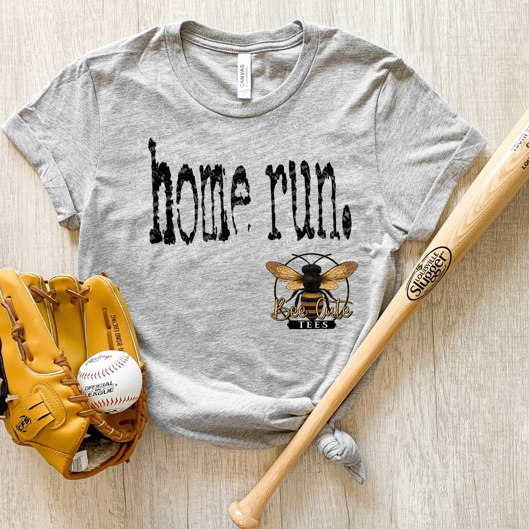 Gray t-shirt with 'home run' and bee logo, baseball glove, ball, and bat on wooden surface